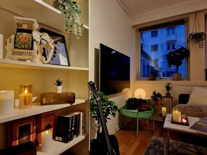 Ein gemütliches Wohnzimmer mit Fernseher, Büchern, einer Gitarre und einem Fenster mit Blick auf Gebäude.