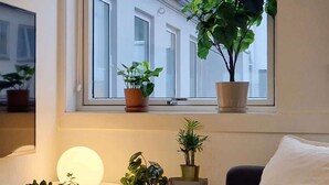 Une pièce chaleureuse avec une grande fenêtre, une petite table d’appoint verte ornée de plantes en pot, une table de chevet avec une bougie et un livre, et un tapis moelleux.