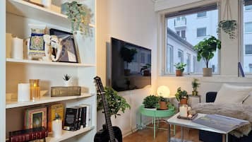 Ein gemütliches Wohnzimmer mit Bücherregal, Gitarre, Fernseher und einem Fenster mit Pflanzen.
