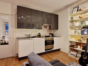 Eine kompakte Küche mit weißen Schränken, einem eingebauten Backofen und einem Essbereich mit einer grauen Couch.