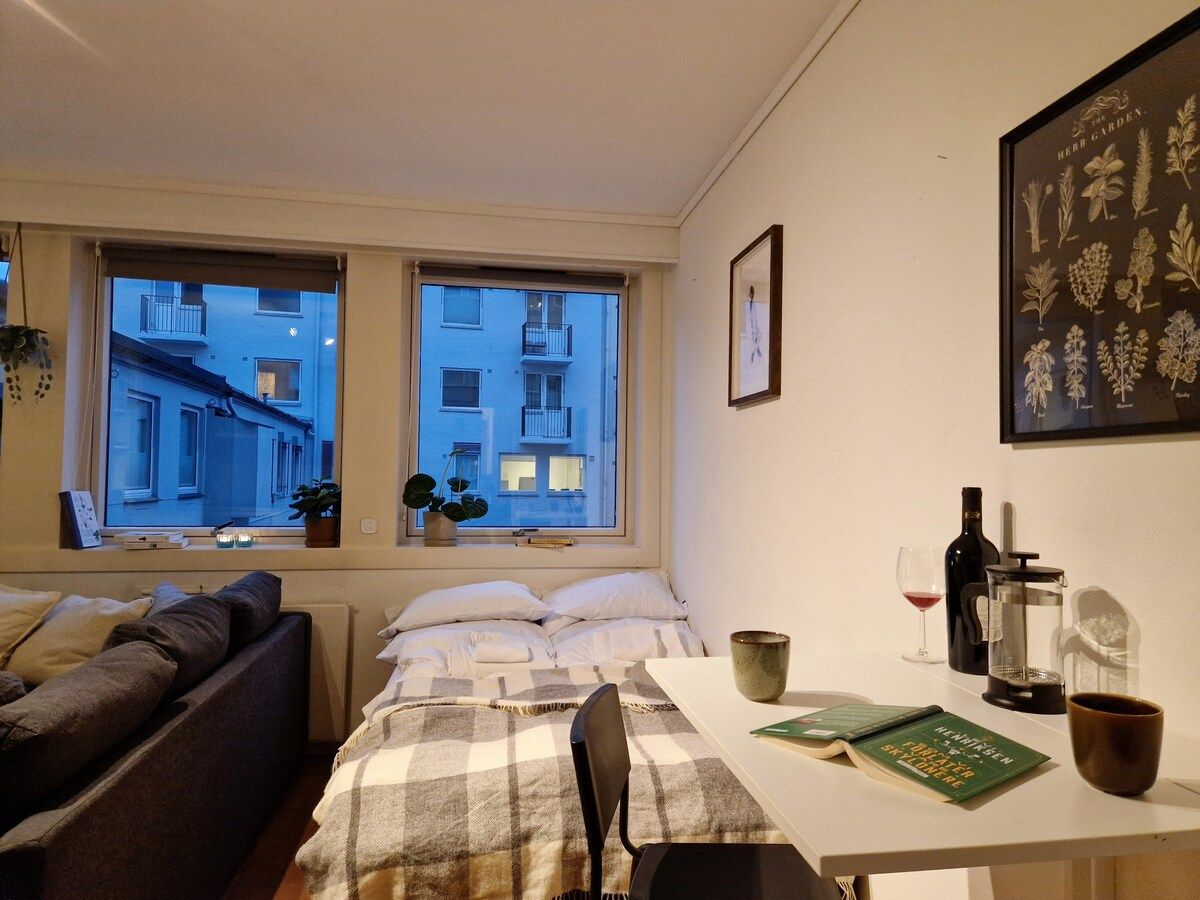 A bedroom with a bed, a window with a view of buildings, a table with a book, a wine bottle, and a coffee maker.