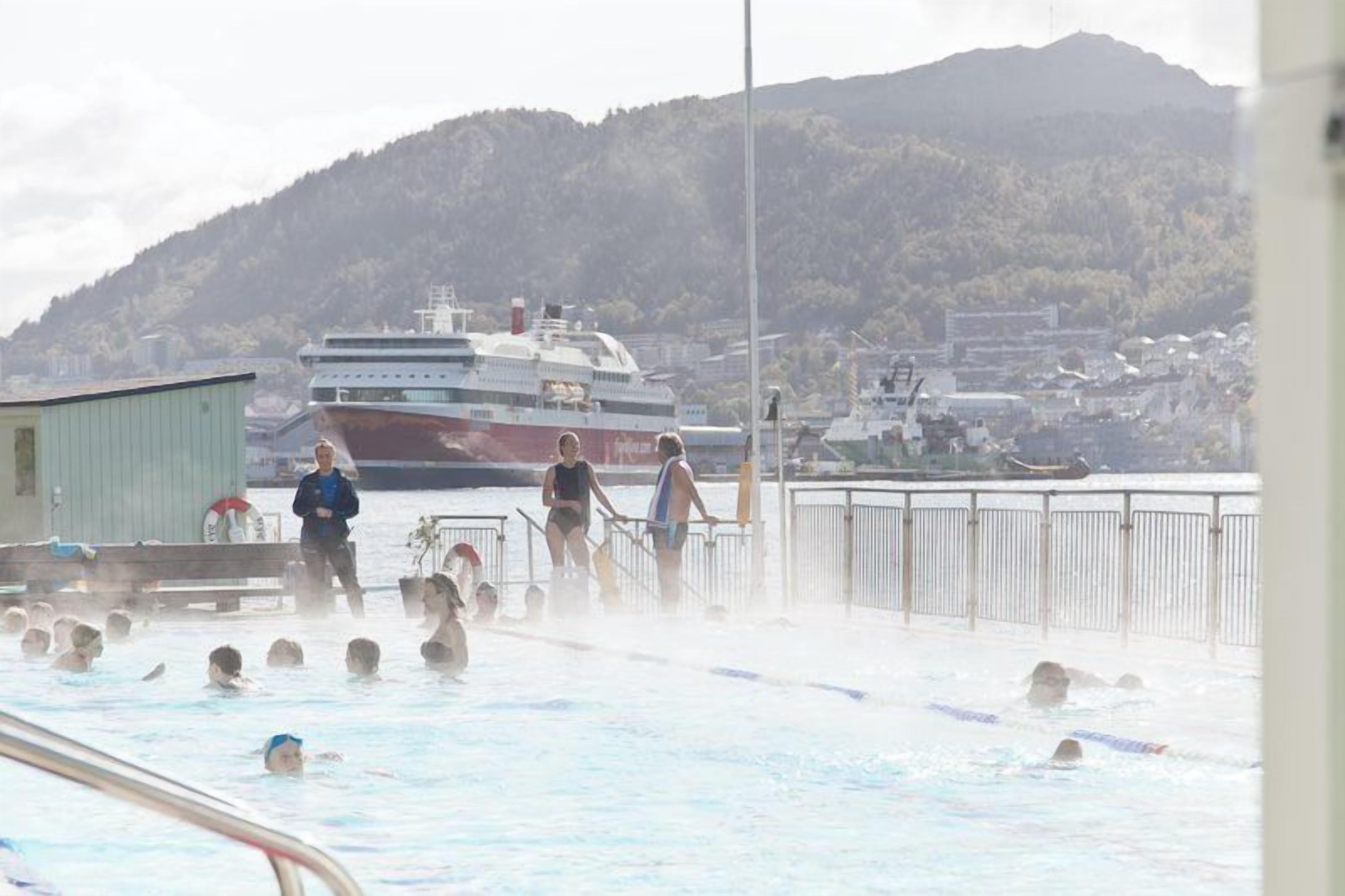 Una piscina con persone a nuotare, una grande nave da crociera sullo sfondo e un paesaggio collinare.