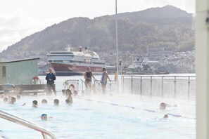 Ein Schwimmbecken mit darin badenden Personen, im Hintergrund ein großes Kreuzfahrtschiff und eine hügelige Landschaft.
