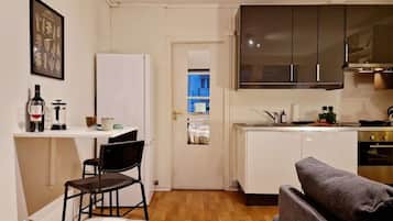Un espacio habitable compacto con kitchenette, una zona de comedor con una silla, un sofá y una puerta que da a una habitación azul.