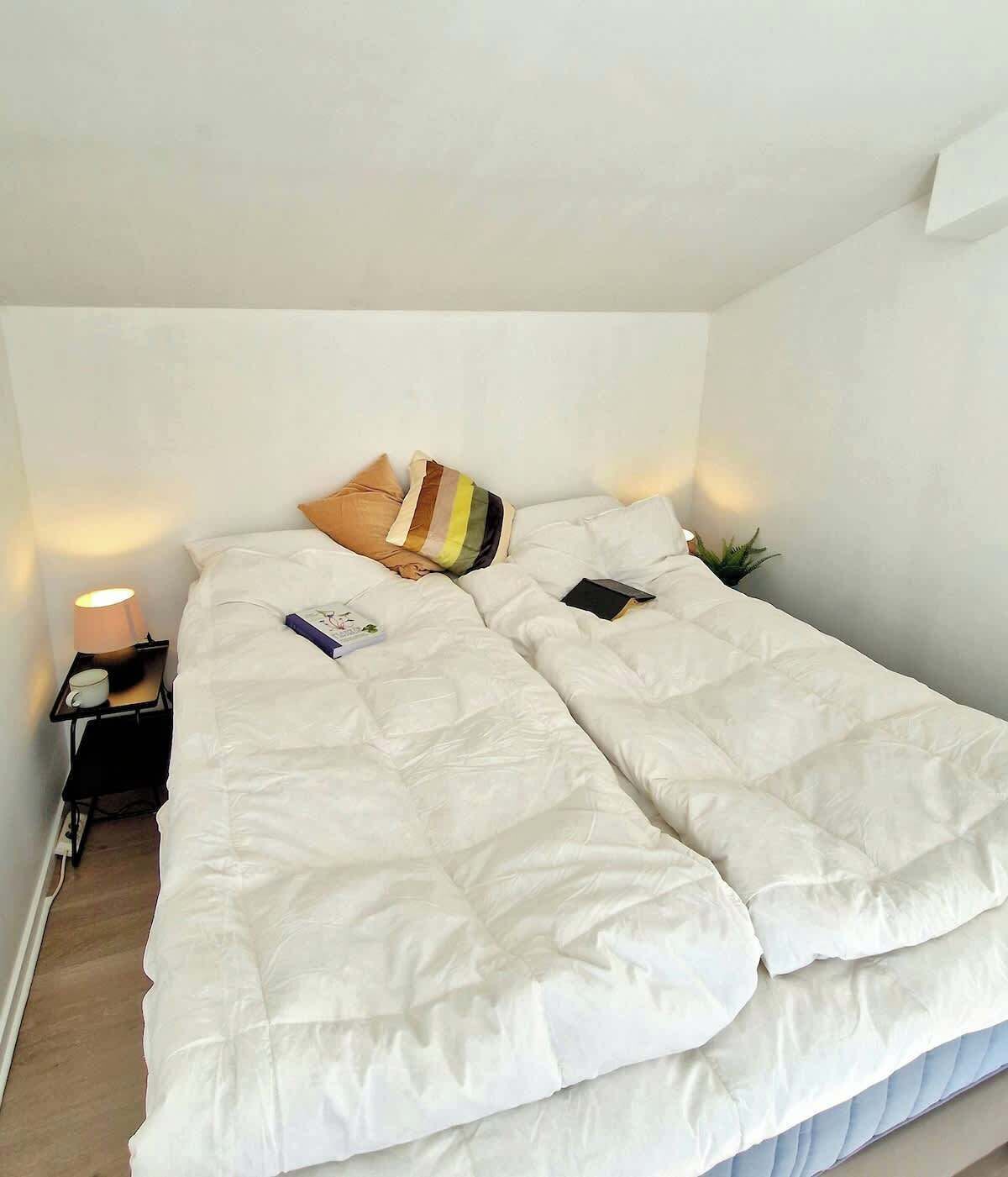 Una cama con ropa de cama blanca, una almohada a rayas y un libro encima. Hay una mesita de noche con una lámpara y una planta pequeña al lado de la cama.