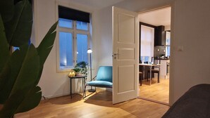 Une pièce avec un plancher en bois, une chaise bleue, une petite table ornée d’une plante et de bougies, et une porte donnant sur un coin cuisine.