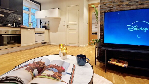 Un salon moderne avec un téléviseur à écran plat affichant le logo Disney, une cuisine avec des meubles blancs et un coin repas avec une table mise pour deux personnes.