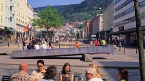 Une place publique où l’on voit des gens assis à des tables, une fontaine et des bâtiments en arrière-plan.