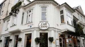 Un bâtiment d’angle avec une enseigne « Café Opera », des chaises en bois à l’extérieur et une entrée bien visible.