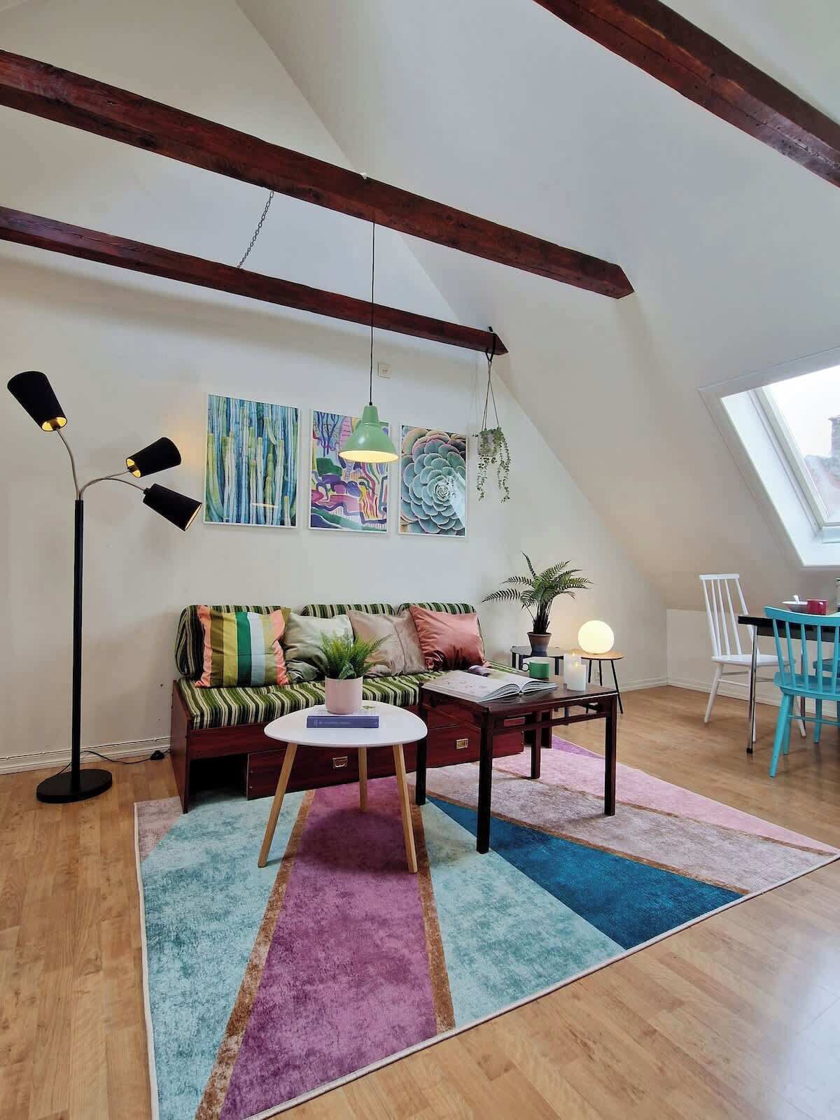 Um apartamento loft aconchegante com teto inclinado, uma viga de madeira, um sofá colorido com listras, uma mesa de centro redonda, uma mesa lateral com abajur e uma área de jantar com uma cadeira azul.
