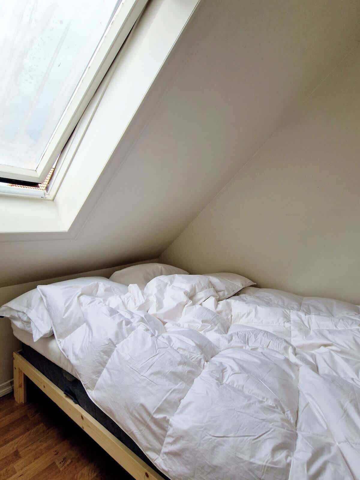 Uma cama de solteiro num quarto com teto inclinado e claraboia.