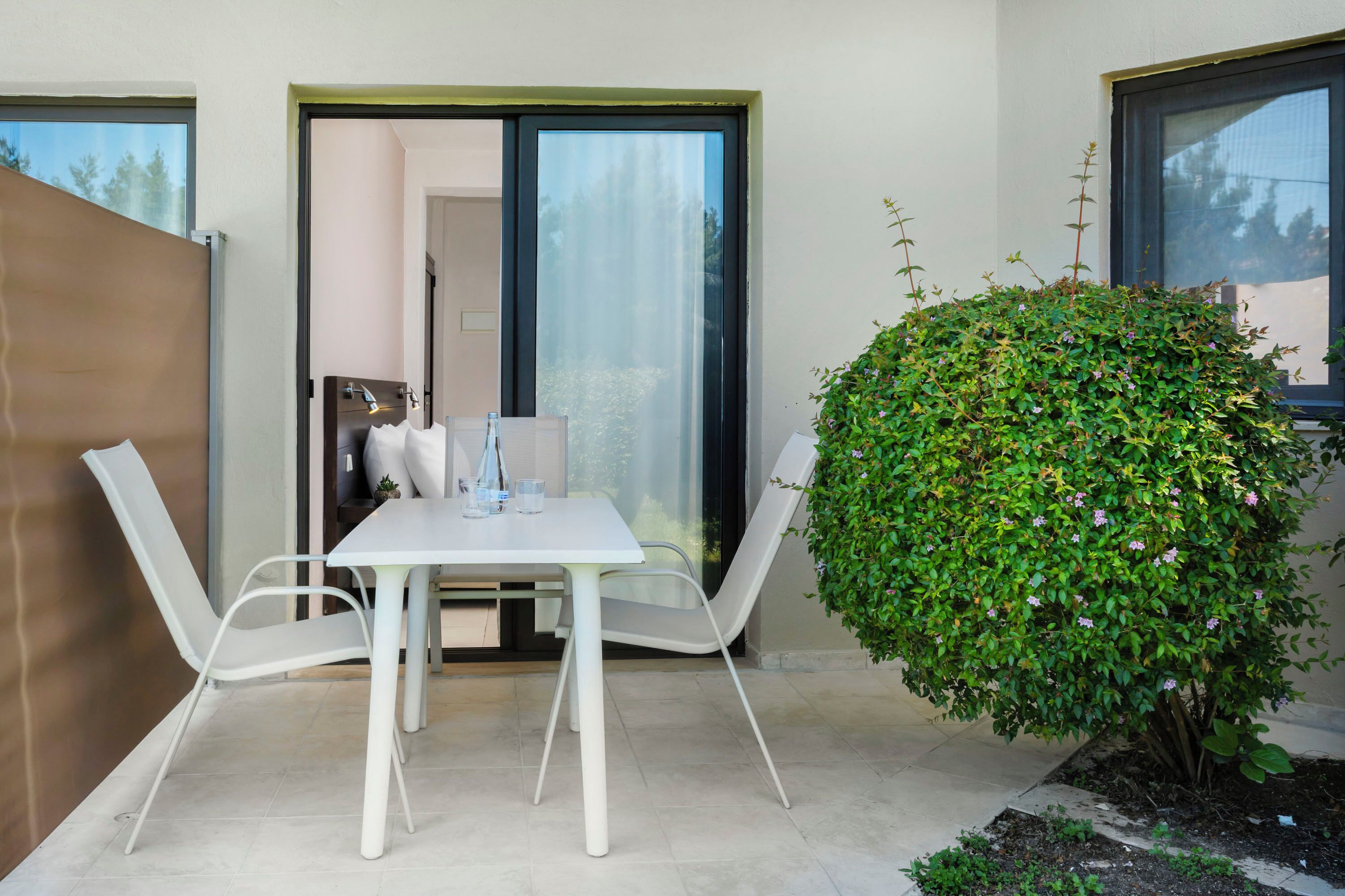 A modern outdoor dining area with a white table and chairs, a large green bush, and a sliding glass door leading to an interior space.