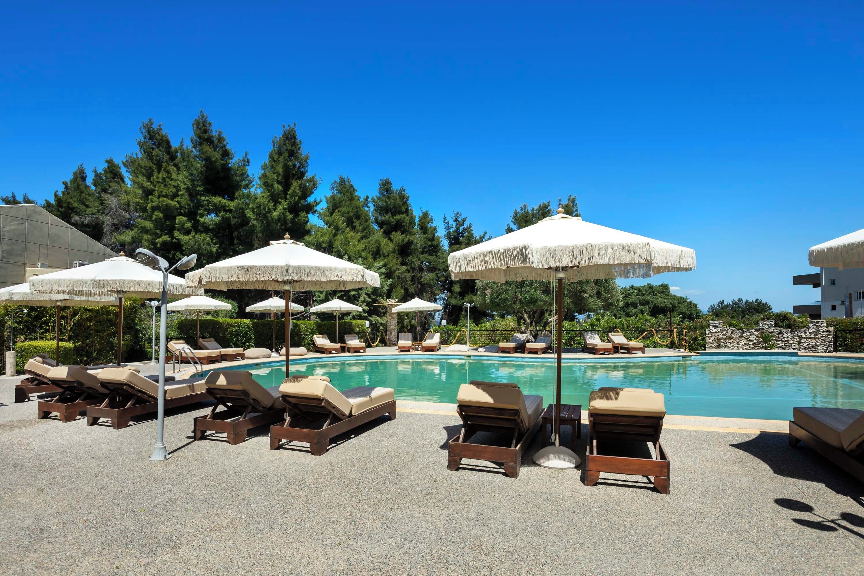 A poolside area with lounge chairs and umbrellas, surrounded by trees and a clear blue sky.