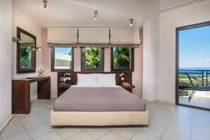 Quarto com cama grande, escrivaninha e mesa de cabeceira. Janelas com cortinas, vista para o exterior e varanda com guarda-corpo.