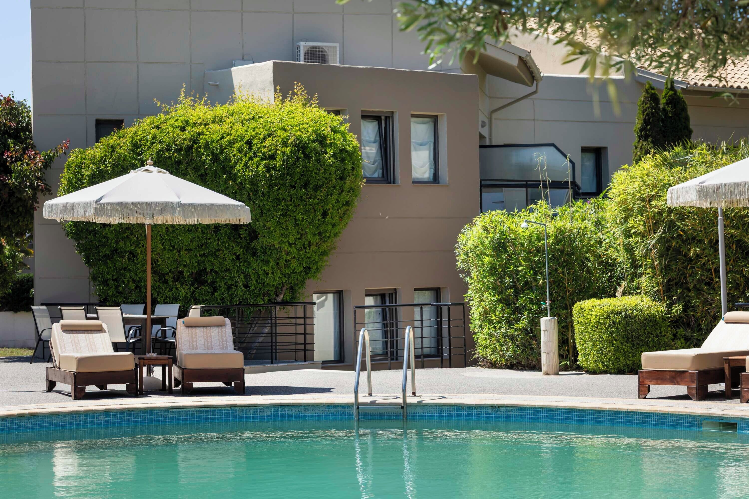 A pool area with lounge chairs and umbrellas, surrounded by greenery and a modern building.