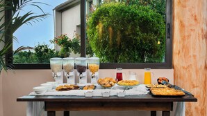 A breakfast buffet with various food items and beverages, set against a window with a view of greenery. - Ohana Hotel (Kassandra)