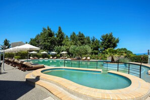 Piscina com espreguiçadeiras e guarda-sóis, cercada por árvores e um céu azul claro.