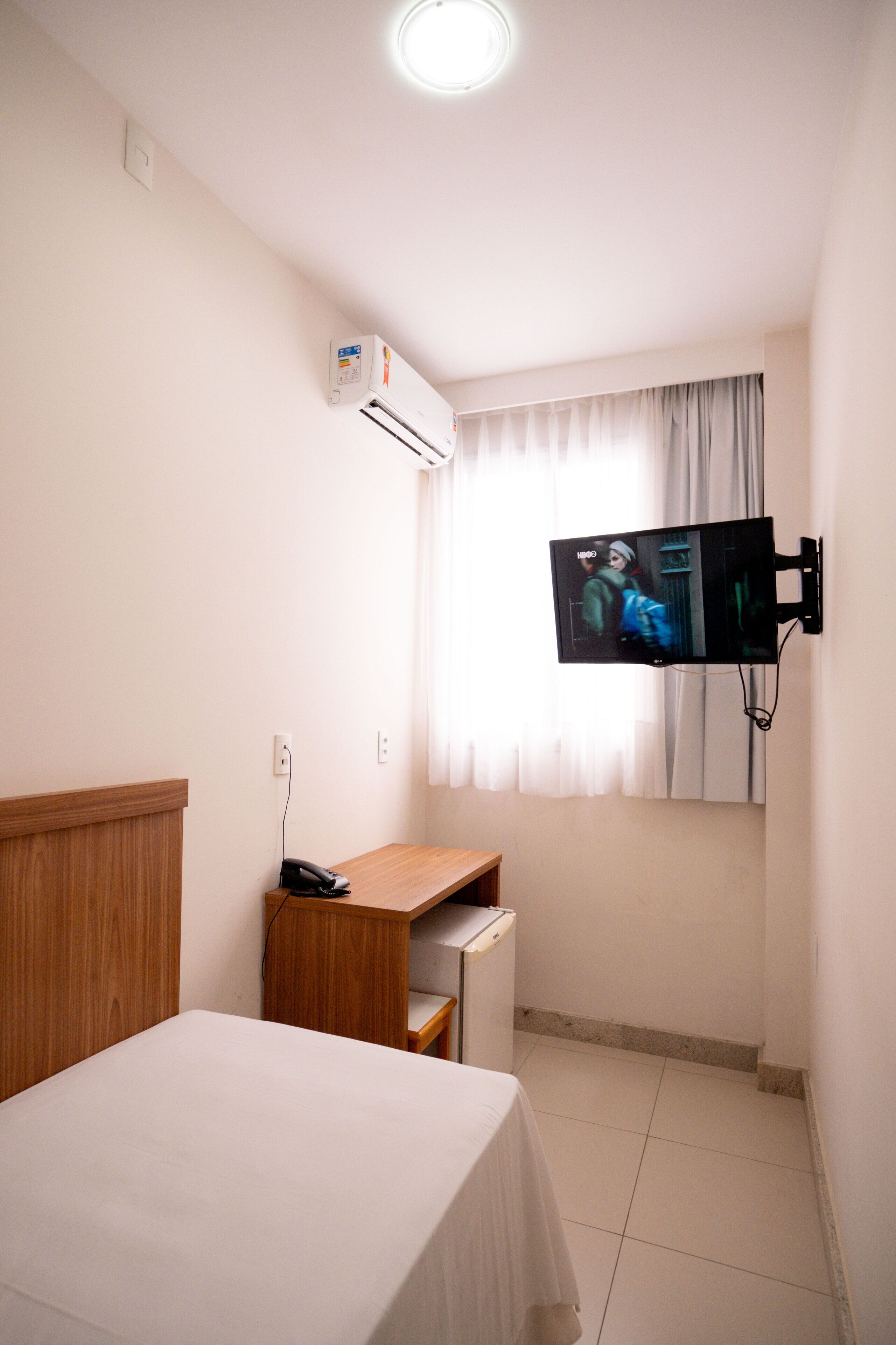 A small hotel room with a bed, a desk, a television mounted on the wall, and a window with curtains.