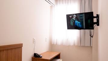 A small hotel room with a bed, a desk, a television mounted on the wall, and a window with curtains.