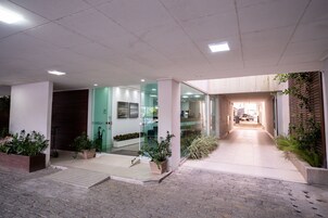 Entrada de edifício com caminho pavimentado, portas de vidro e plantas em vasos.