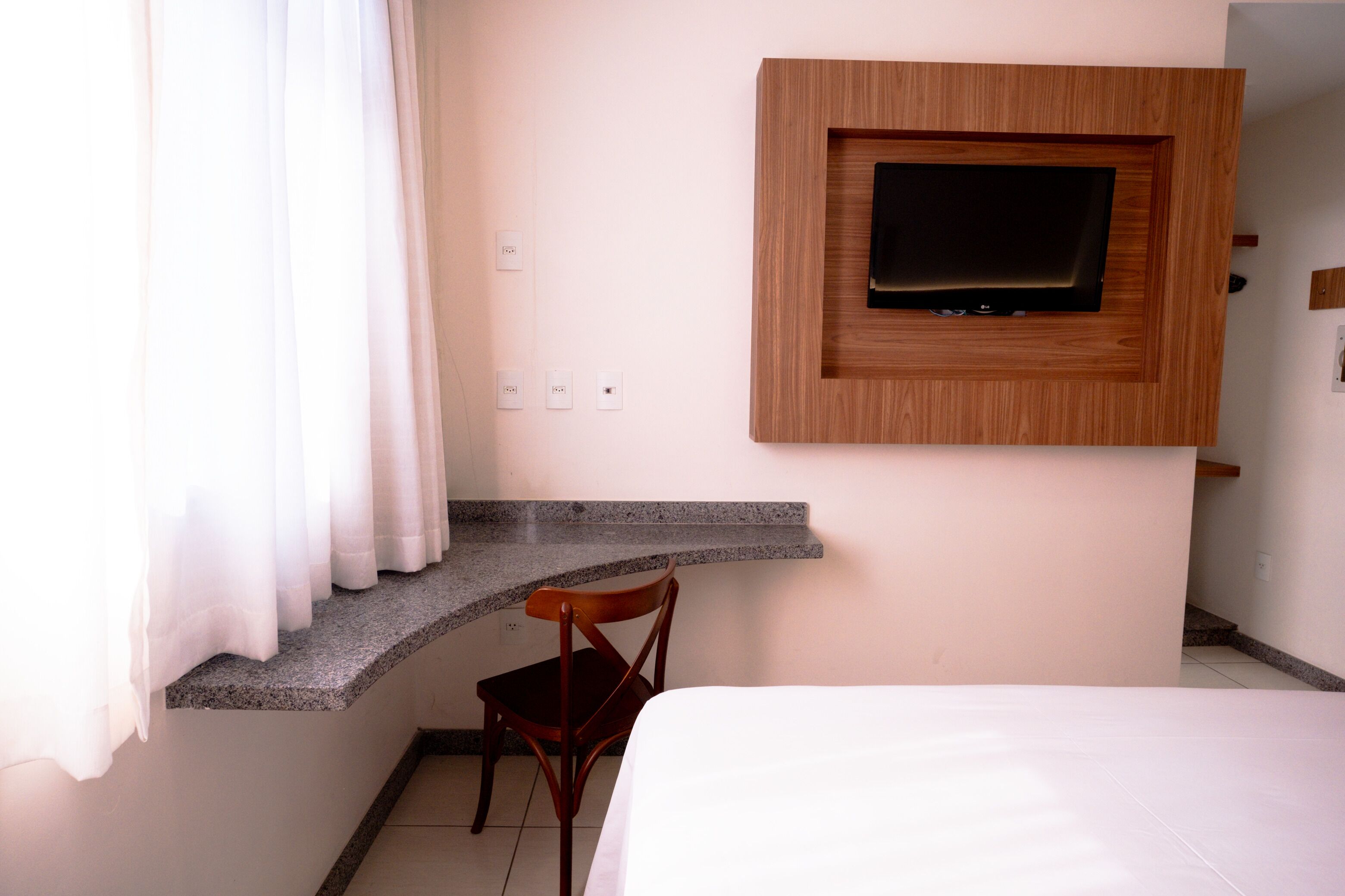 A hotel room with a bed, a TV mounted on a wooden panel, a chair, and a small desk area.