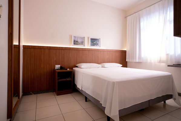 A hotel room with a wooden headboard, a bed with white linens, a nightstand, and two framed pictures on the wall. - Hotel Conceição (Linhares)