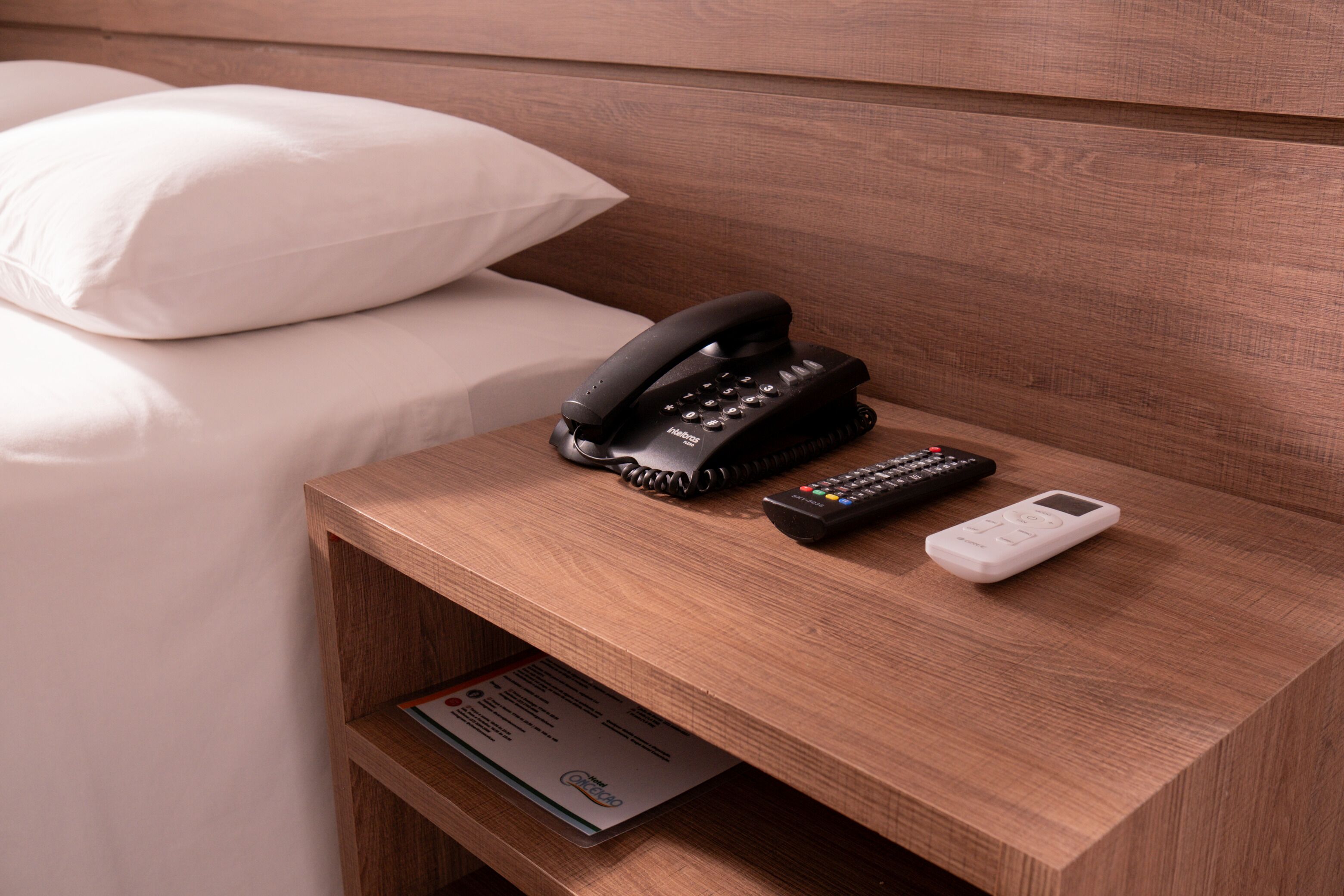 A wooden bedside table with a black telephone, a remote control, and a white device, likely a thermostat or control panel, in a hotel room.