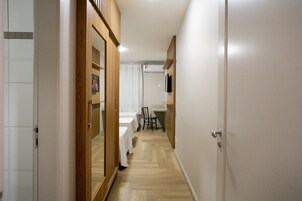 Um corredor estreito com uma porta de madeira à esquerda, uma parede branca com uma TV montada à direita e uma cama com colcha branca no centro.