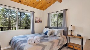 A bedroom with a large bed, two windows with curtains, a nightstand with a lamp, and a picture of a deer on the wall.
