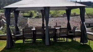 Un espace extérieur abrité, aménagé avec une table et des chaises, dont le décor est constitué d’un ciel nuageux et d’une vue lointaine sur un bâtiment.