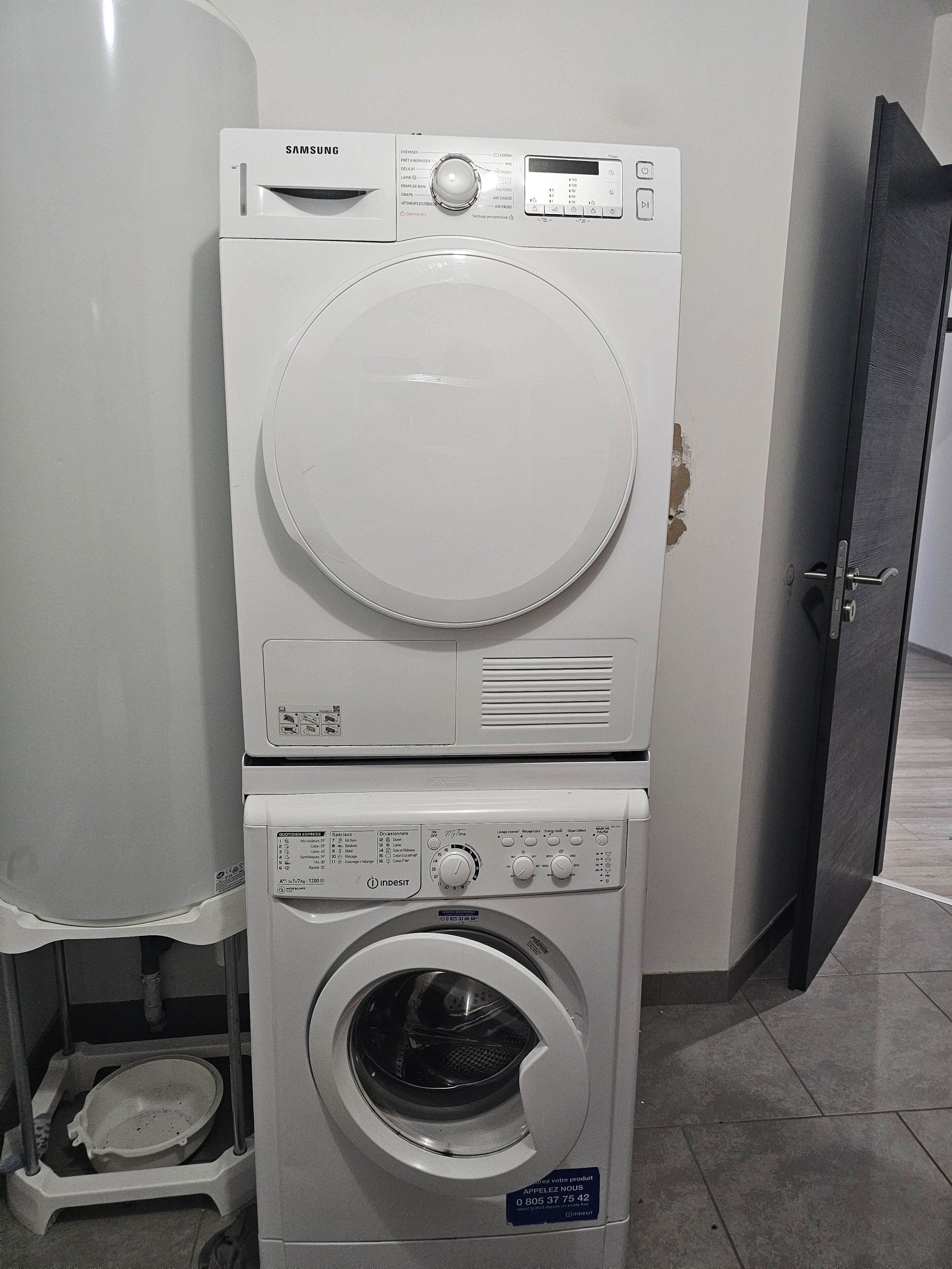 Una rentadora i una màquina d'assecar roba apilades en una habitació amb terra de rajola i una porta al fons.