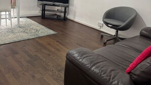 A living room with a brown leather sofa, a grey office chair, a flat-screen TV on a stand, and a white chair with a red cushion. - Calm and Cosy Apartment in Central London (London)
