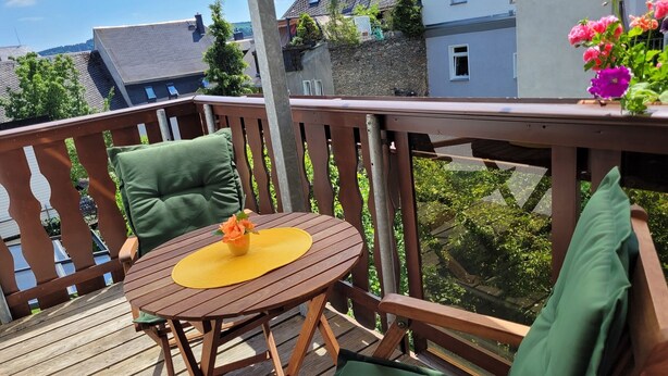 Eine Holzterrasse mit rundem Tisch und zwei Stühlen, einem kleinen Topfpflanzen und Blick auf benachbarte Häuser.
