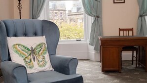 A room with a blue armchair, a wooden dining table, and a window with curtains. - Location! Location! Location! A Golf Lover’s Dream (St Andrews)