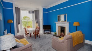 A living room with blue walls, a fireplace, a dining table with chairs, a sofa, and a TV. - Location! Location! Location! A Golf Lover’s Dream (St Andrews)