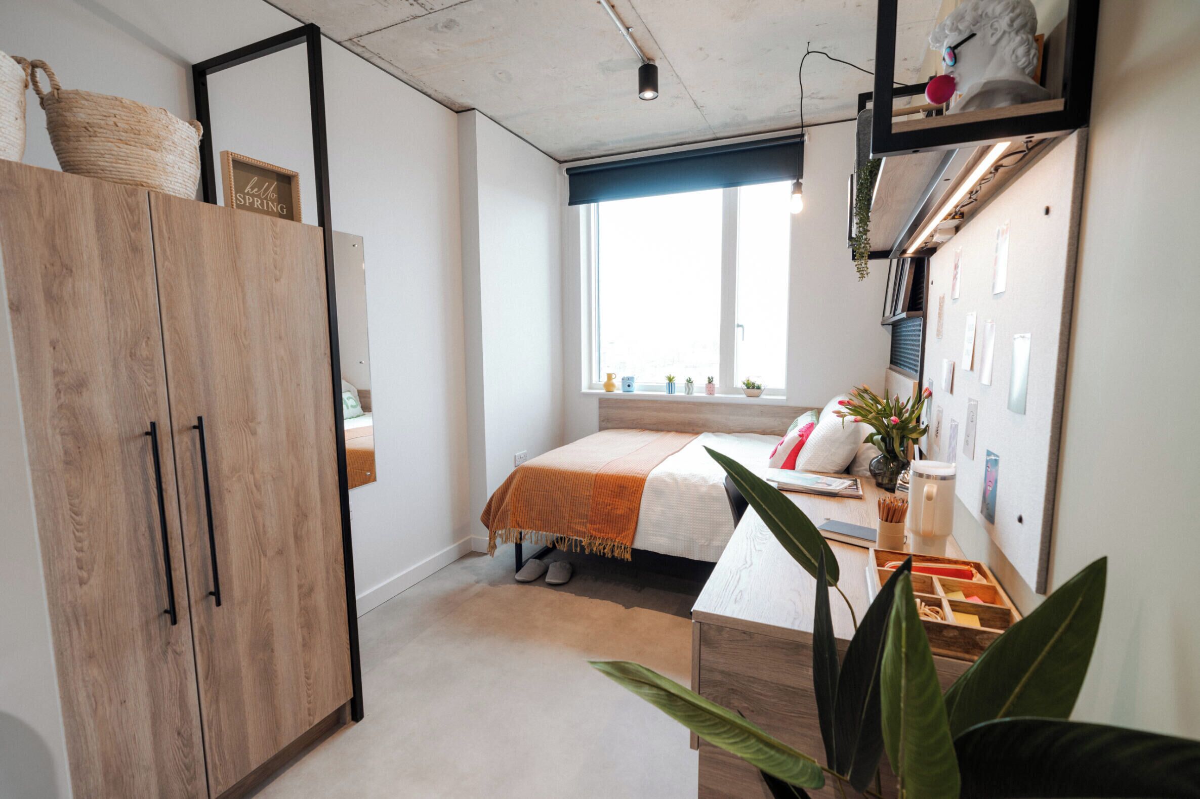 A bedroom with a bed, a wooden wardrobe, a window with plants, and a desk with a plant.