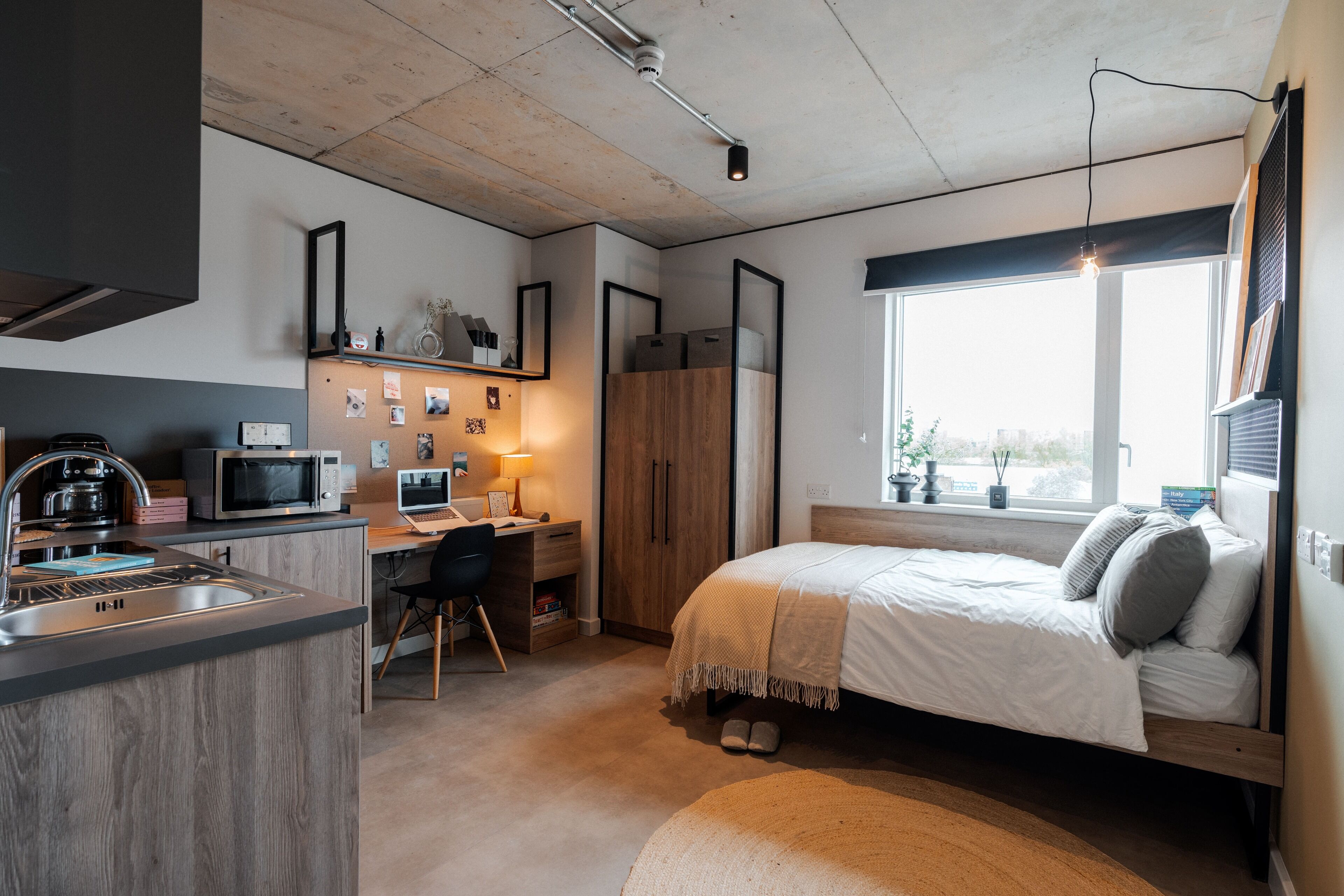 A compact studio apartment with a kitchenette, a bed, a desk, and a large window providing natural light.