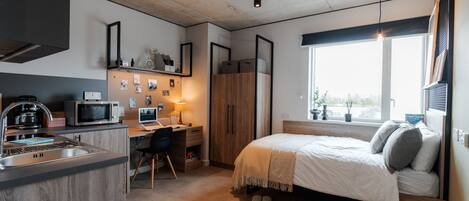 A compact studio apartment with a kitchenette, a bed, a desk, and a large window providing natural light.