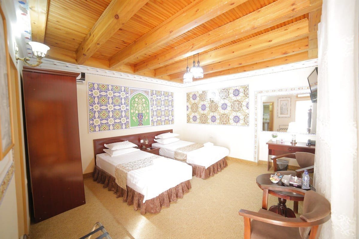 A room with two beds, wooden ceiling beams, patterned wall tiles, a desk with a chair, and a mirror.