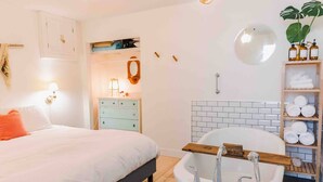 A bedroom with a bed, a dresser, a vanity, and a bathroom with a bathtub and shelves.