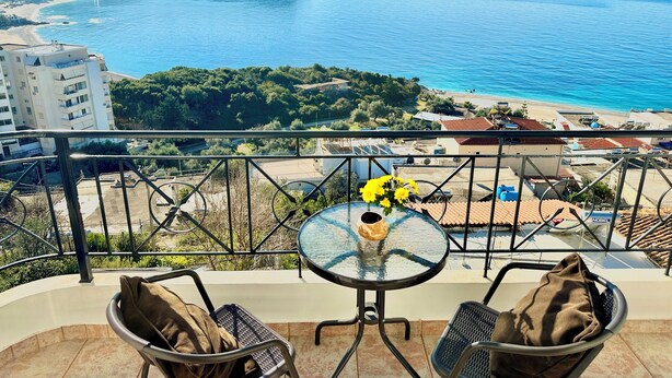 A balcony with a glass table and chairs, overlooking a coastal view with buildings and a hill.
