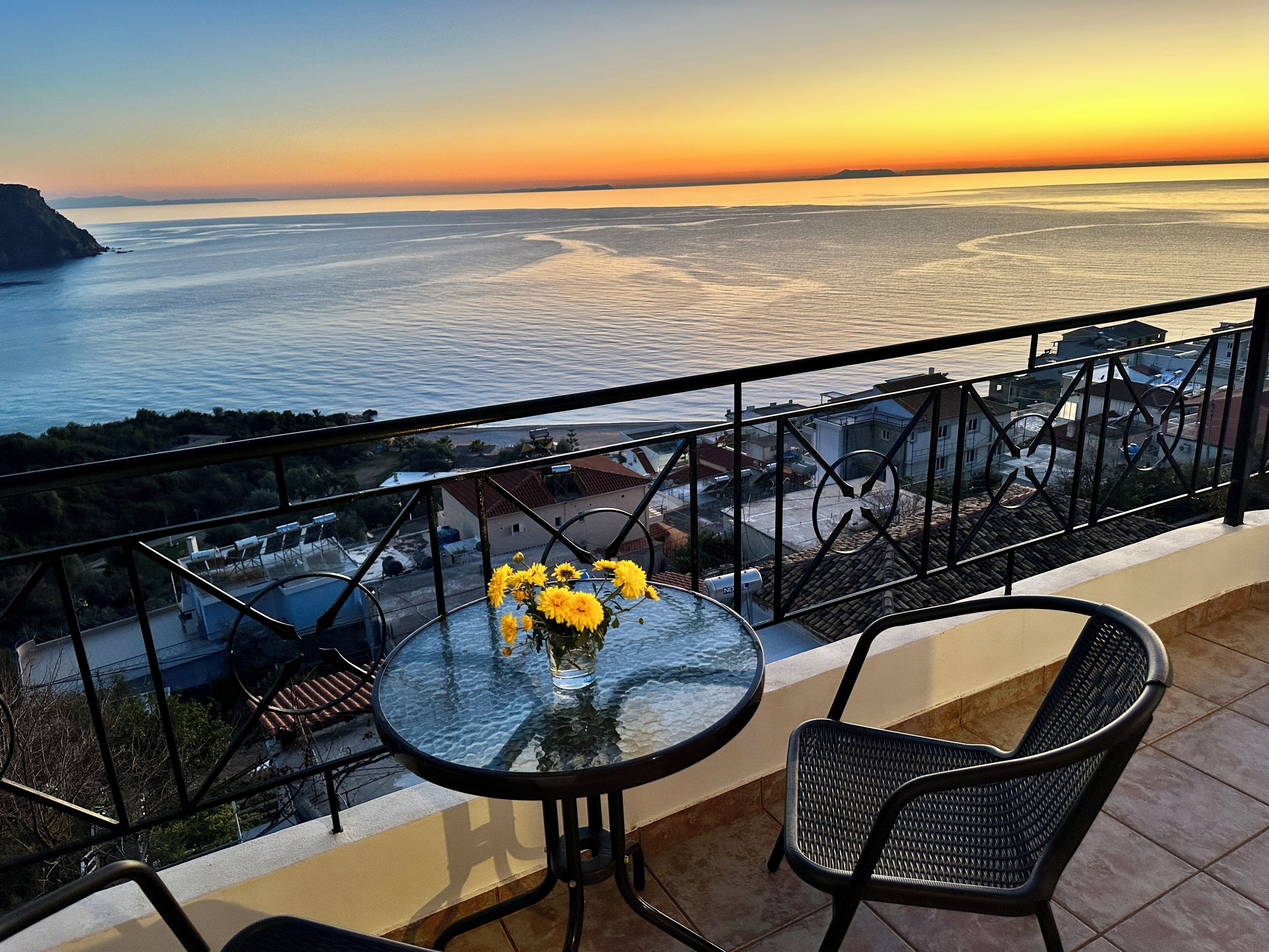 A balcony with a glass table and chairs, overlooking a coastal view with a sunset.