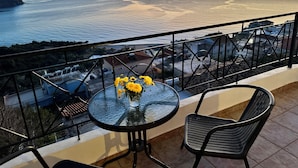 A balcony with a glass table and chairs, overlooking a coastal view with a sunset.