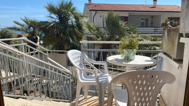 A balcony with white plastic chairs and a table, overlooking a building with a red-tiled roof and palm trees. - Apartment with access to the Costa dei Trabocchi bike path (ortona)