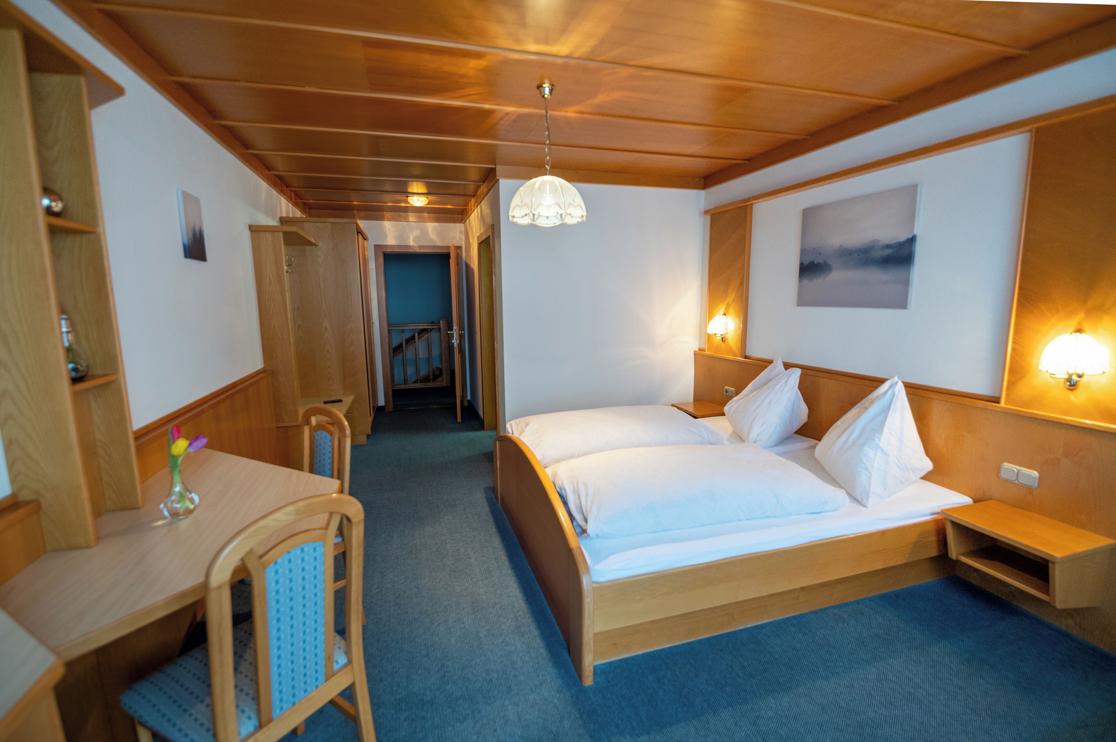 A hotel room with a wooden bed, a desk, and a chair. There is a hallway leading to another room.