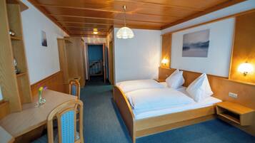 Habitación de hotel con una cama de madera, un escritorio y una silla. Se observa un pasillo que conduce a otra habitación.
