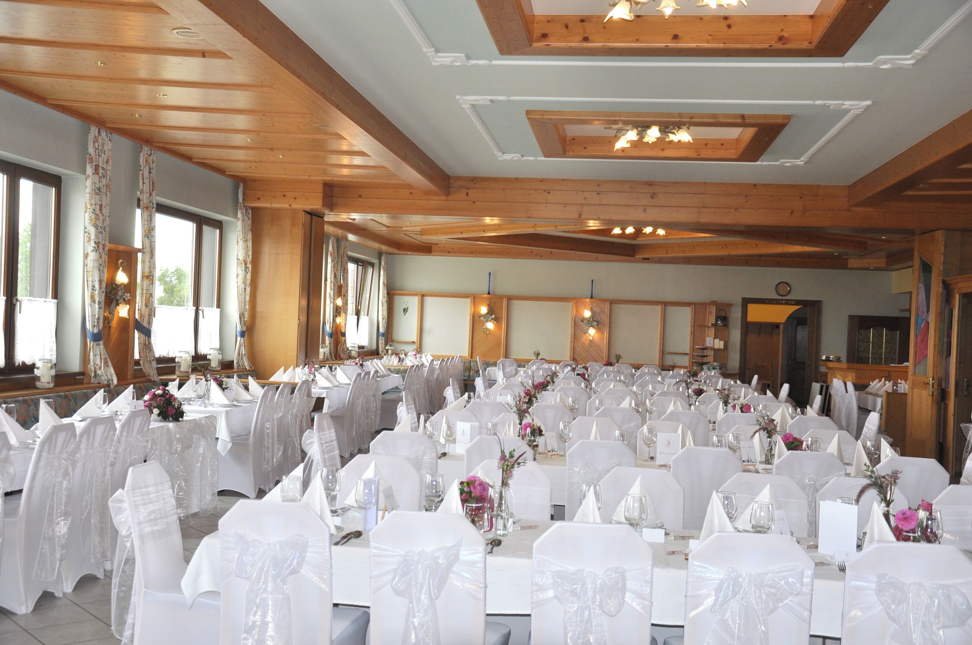 A banquet hall with round tables covered in white tablecloths and chairs, wooden beams on the ceiling, and large windows.