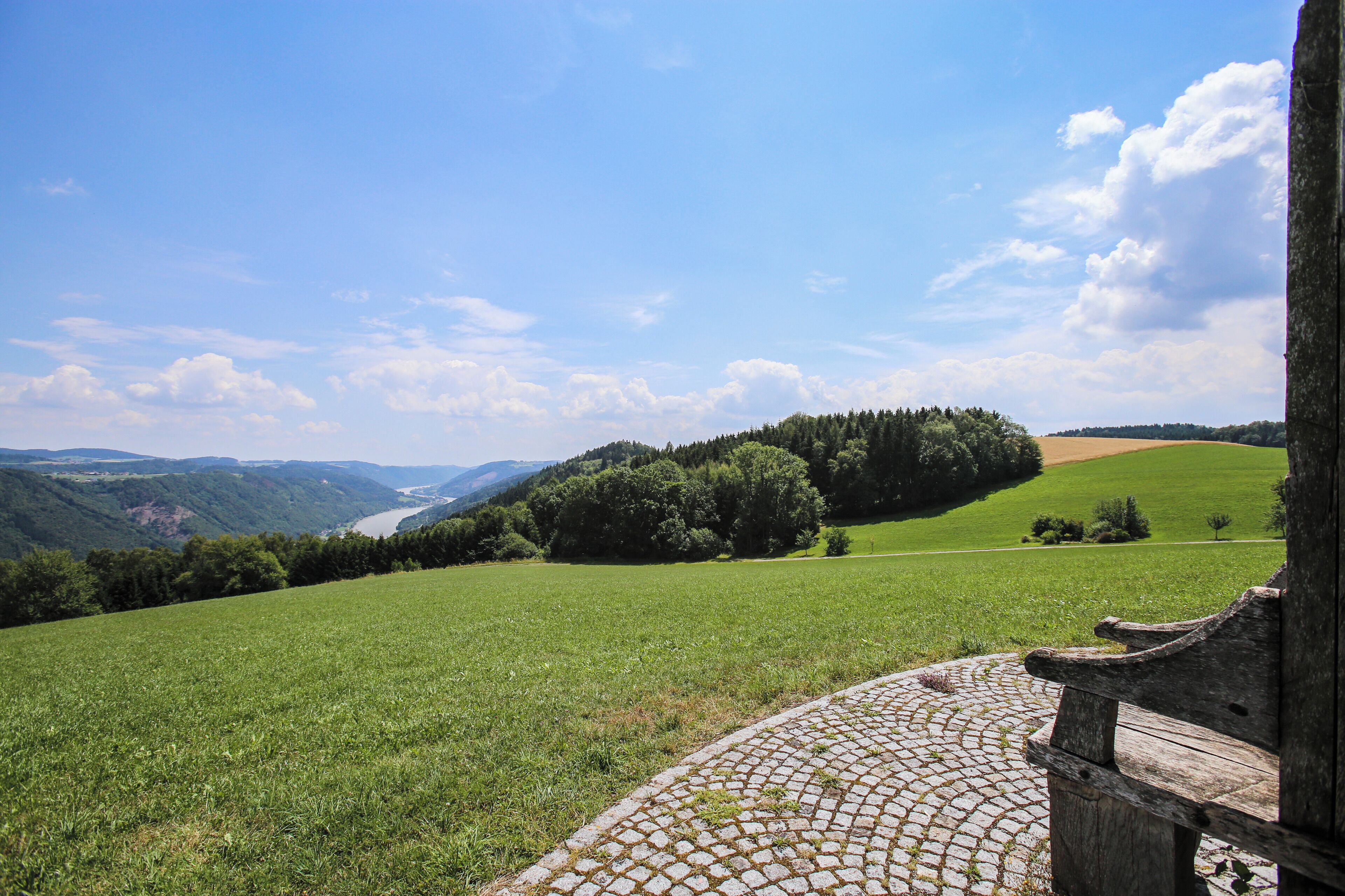A cobblestone path leading to a wooden bench overlooking a green field and forested hills.