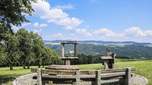 A stone bench and table set in a circular stone pattern, surrounded by grass and trees, with a view of distant hills and a clear sky. - Kirchenwirt St. Aegidi (St. Aegidi)