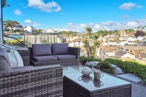 A balcony with wicker furniture, a glass table, and a view of a town with buildings and a church. - 2 Bed in Brixham (oc-p30779) (Brixham)